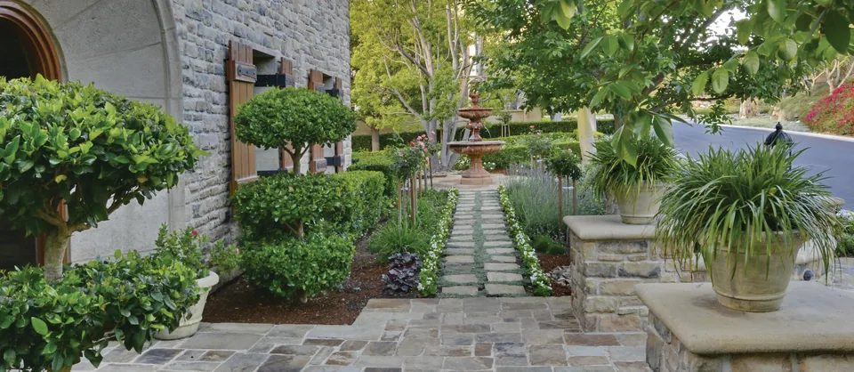 A flagstone path through a shaded courtyard garden of clipped shrubs, potted plants and trees beside a stone house.